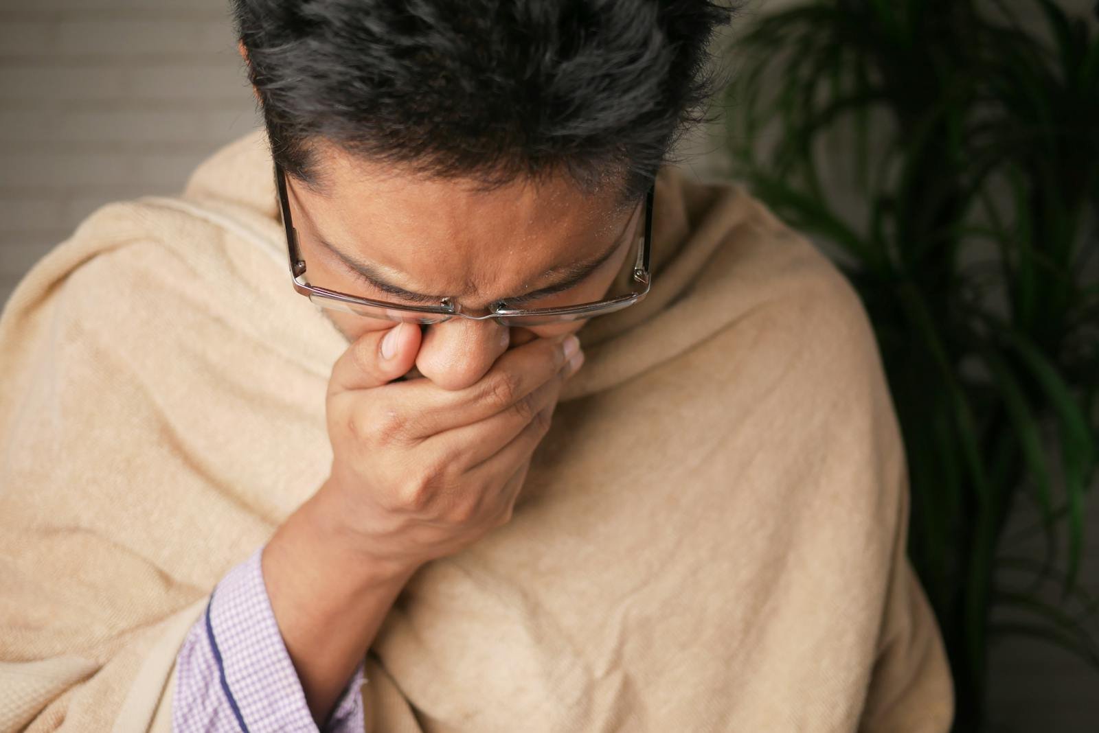 Adult wearing glasses coughs into hand indoors, indicating illness or flu symptoms.